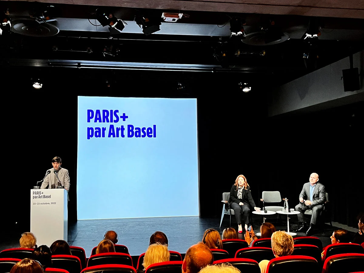 A young French white man stands at a podium with two microphones. To his right, is a white woman and white man seated on the stage. Behind is a large screen reading 'PARIS+ par Art Basel.'